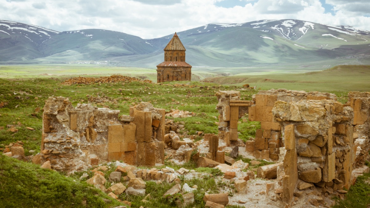 The ruined city of Ani: a lone red-stone church with a conical dome standing alone on open grassland, tumbled masonry in the foreground, snow-streaked mountains behind.