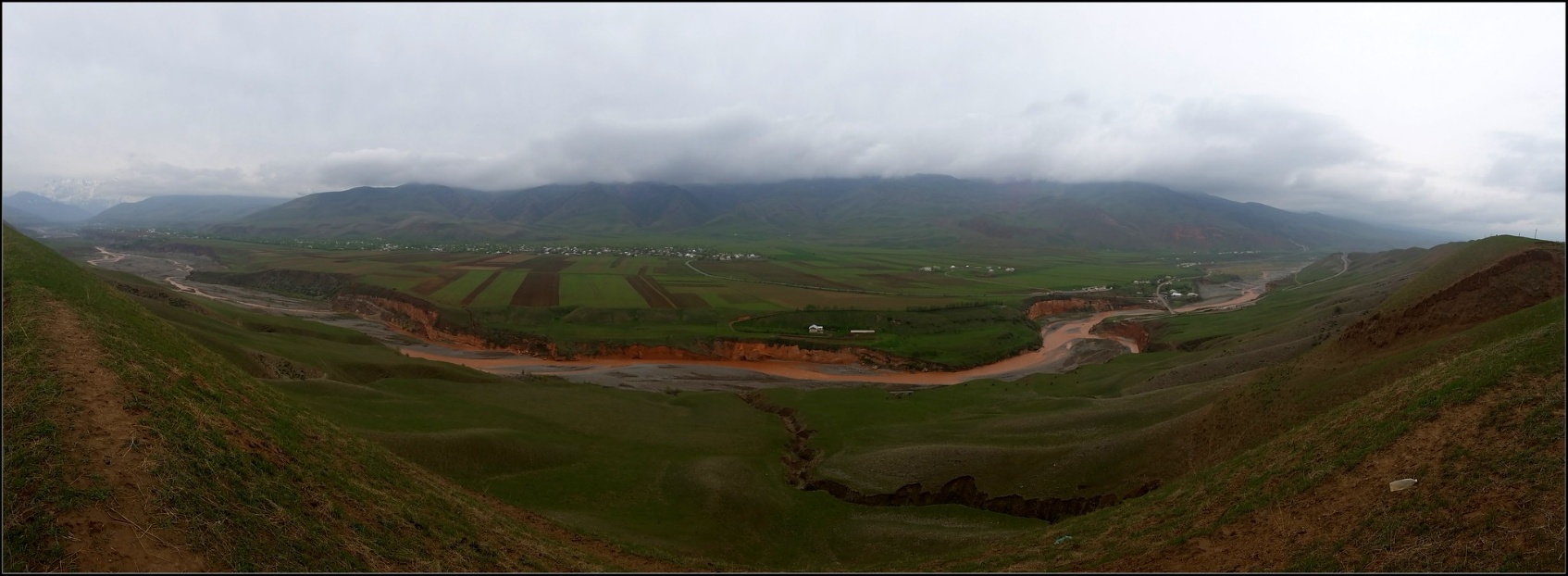 A vast, misty landscape with a river flowing through undulating green fields, flanked by mountains under a cloudy sky.